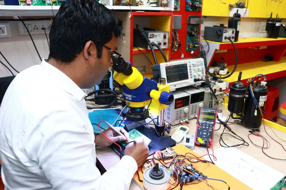 An electronics technician inspecting a circuit board under a stereo microscope, with an oscilloscope and bench power supply alongside