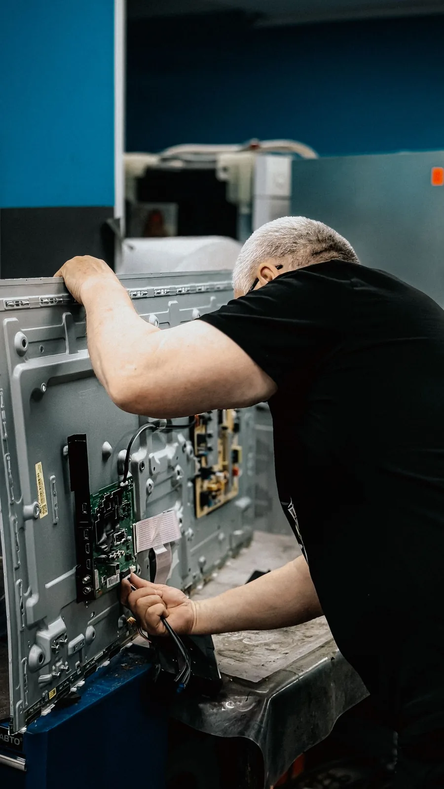A technician working on the exposed back of a flat-screen television, with the mainboard, power board and ribbon cables visible